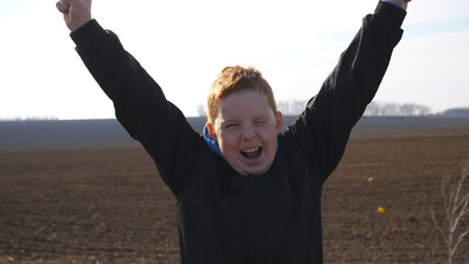 Little red-haired boy with freckles looks into camera and raises hands rejoicing achievement standing on ploughed meadow. Portrait of happy male child with positive emotion on his face at plowed field