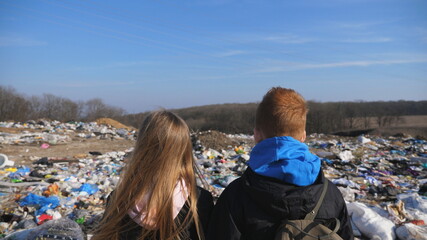 Rear view of young girl and boy stands against the blurred background of dirty garbage dump. Little...