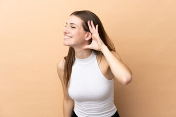 Young caucasian woman isolated on beige background listening to something by putting hand on the ear