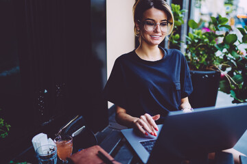 Cheerful student in optical eyewear watching webinar on laptop computer sitting in coffee shop...