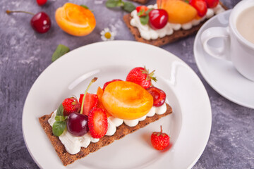 Crisp fitness health bread with creme cheese, fruit and berries and cup of tea on the white plate on the white wooden background.