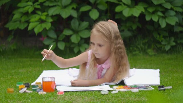 Blond girl with wavy hair lies on a green lawn and paints a picture
