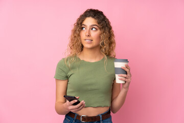 Young blonde woman with curly hair isolated on pink background holding coffee to take away and a mobile while thinking something