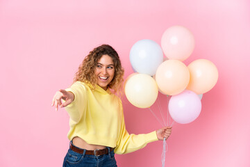 Young blonde woman with curly hair catching many balloons isolated on pink background pointing finger to the side