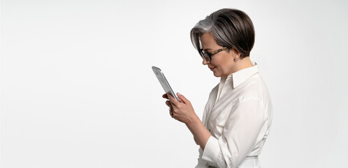 Fototapeta premium Profile view of serious woman working with digital tablet. Cheerful Mature business woman using tablet computer standing on white background. Horizontal template for ad banner with textspace at left.