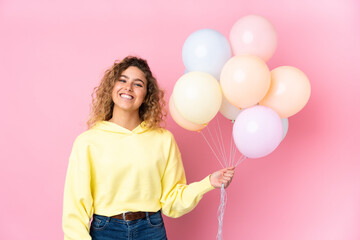 Young blonde woman with curly hair catching many balloons isolated on pink background applauding