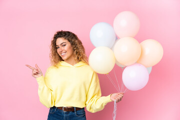 Young blonde woman with curly hair catching many balloons isolated on pink background pointing finger to the side