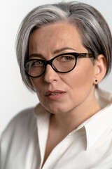 Fototapeta premium Smart mature woman in white shirt looking thoughtfully at the camera. Pretty Caucasian gray haired model in glasses posing in studio. Close up studio portrait.