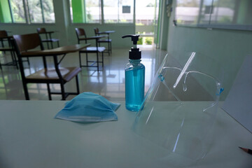 Medical mask and alcohol hand wash gel on the teacher 's desk in front of the classroom with the face shield in the empty classroom. Concept during the Coronavirus disease COVID-19 outbreak.