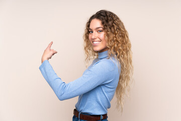 Young blonde woman with curly hair wearing a turtleneck sweater isolated on beige background pointing back