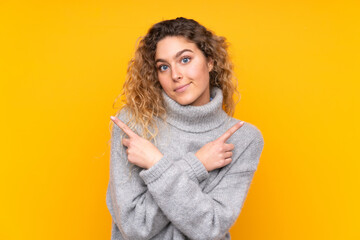 Young blonde woman with curly hair wearing a turtleneck sweater isolated on yellow background pointing to the laterals having doubts