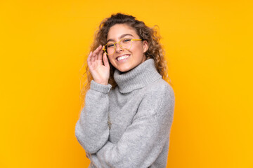 Young blonde woman with curly hair wearing a turtleneck sweater isolated on yellow background with...