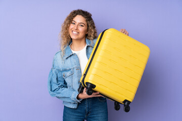 Young blonde woman with curly hair isolated on purple background in vacation with travel suitcase...