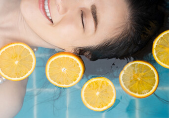 Asian woman is swimming with a slice of orange floating above the water in a blue pool and enjoying...