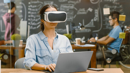 Young Female Creative Developer Wearing Virtual Reality Headset  Works on a Laptop, She Develops New AR, VR Games and Applications. In the Background Stylish Office with Talented People.