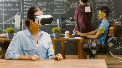 Young Female Creative Developer Wearing Virtual Reality Headset  Works on a Laptop, She Develops New AR, VR Games and Applications. In the Background Stylish Office with Talented People.