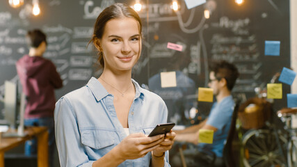 Charming Young Woman Uses Smartphone, Smiles Beautifully on Camera While Standing in the Busy Creative Office with Stylish Talented People. - obrazy, fototapety, plakaty