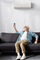 Senior man holding remote controller while sitting on sofa under air conditioner