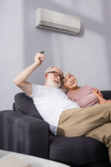 Selective focus of smiling elderly man embracing wife and using remote controller of air conditioner at home