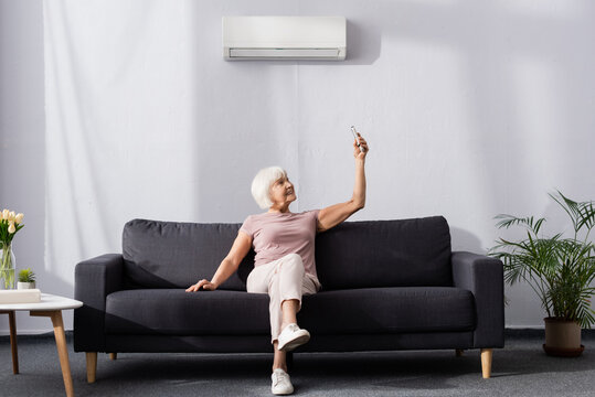 Smiling senior woman switching air conditioner with remote controller on couch