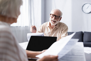 Selective focus of smiling man looking at wife with papers near laptop at home