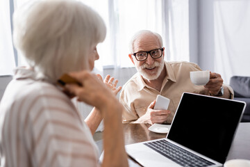 Selective focus of smiling senior man holding cup and smartphone near wife with credit card pointing with hand and laptop on table