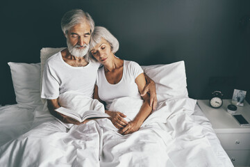 Happy family. Cozy home. Beautiful senior woman and her husband reading book together in bedroom.
