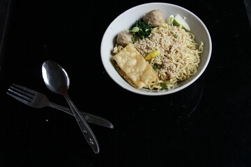 food in a bowl and cutlery with isolated black background. Bakmi or Yahun are food made from noodles with a mixture of meatballs, shredded, chips, pickles and salty broth.