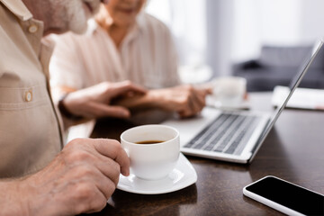 Fototapeta na wymiar Cropped view of senior man holding cup of coffee near digital devices and wife at home