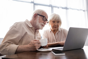 Selective focus of senior man drinking coffee while wife using laptop near smartphone on table
