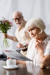 Selective focus of sad senior woman reading news near positive husband with bouquet at home