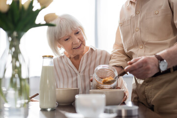 Selective focus of smiling senior woman sitting near man pouring cereals in bowl during breakfast