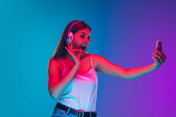 Selfie, listening to music in headphones. Caucasian young woman's portrait on gradient background in neon light. Beautiful model. Concept of human emotions, facial expression, sales, ad. Copyspace.