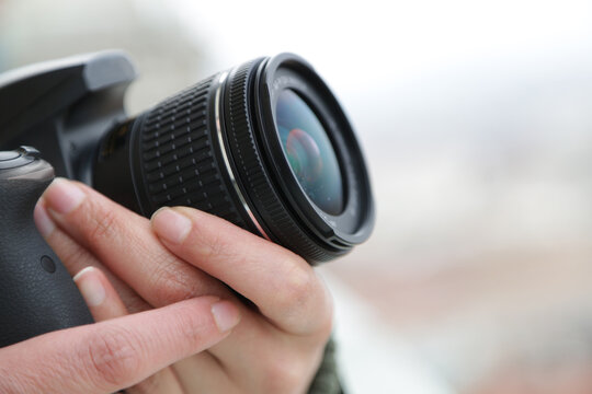A hand holding a modern digital camera. The feminine slim fingers firmly grab the lens of the black DSLR. The shiny glass filter reflects the bright light. A concept of dreamy urban photograph taking.