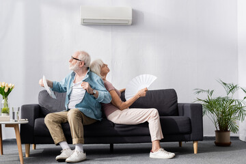 Senior man holding newspaper near tired wife with fan under air conditioner at home