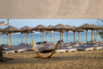 Focus on the wooden board that says beach bar. Behind are umbrellas and sunbeds and blue sea and blue sky. Copy space.