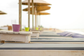 Focus on the cup on the wooden table next to the sun lounger on the beach bar. The beach is empty and there is no one.