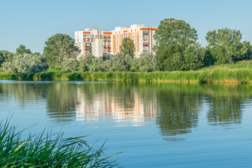 Fototapeta na wymiar Summer landscape in the Zhuravlevsky Godropark with a view of new houses and trees near the Kharkov river and reflection