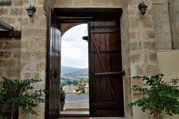Doorway in stone wall. One half of the wooden door is open. Visible pavement, decorative flowers. In the distance, in the valley is a village. On a cloudy sky silhouettes of mountains. Cappadocia 