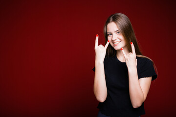 A beautiful European girl in a black T-shirt smiles and shows with her hands a rocker sign on a red background.