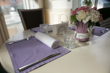 Dining table with cute flower decor and purple napkins in cozy interior across from the window. Anniversary or event concept. Beautiful table with flowers and napkins.
