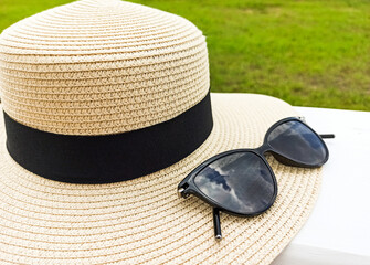 A straw female hat with sun glasses lies on a white table. Hat with a black ribbon and black women's glasses on a background of green grass. Holidays on the coast.