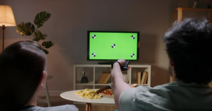 Couple spending time together in the evening, sitting on couch and watching tv with green chroma-key green screen. A template for your clips 4k footage