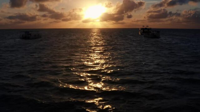 Beautiful aerial tilt up drone shot of an ocean sunrise at Well Beach near Joao Pessoa on a warm summer morning with the water below, golden clouds on the skyline, and small boats in the water.