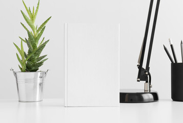 White book mockup with a aloe vera in a pot, lamp and workspace accessories on a white table.