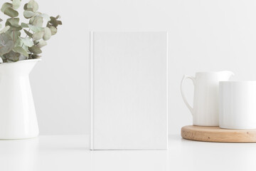 White book mockup with a eucalyptus in a pot and a mug on a white table.