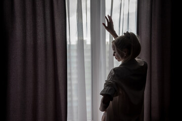 Fototapeta na wymiar Silhouette of girl in white men shirt at window. Portrait of nice female. Home comfort. Slavic woman is standing and touches curtain of tulle. Emotions and relax. Concept image