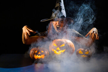 An ominous witch in a hat conjures over a jack-o-lantern. Traditional halloween characters. Mystical fog creeps over pumpkins with carved terrible faces. Woman in carnival costume.