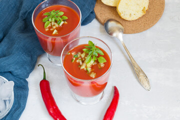 Summer cold tomato gazpacho soup in glass glasses with chopped cucumbers, peppers and greens on a white background. Sliced bread for snacks. Ice for cooling. Vegetarian food