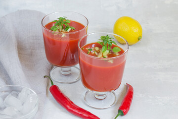 Summer cold tomato gazpacho soup in glass glasses with chopped cucumbers, peppers, lemon and greens on a white background. Ice for cooling. Vegetarian food